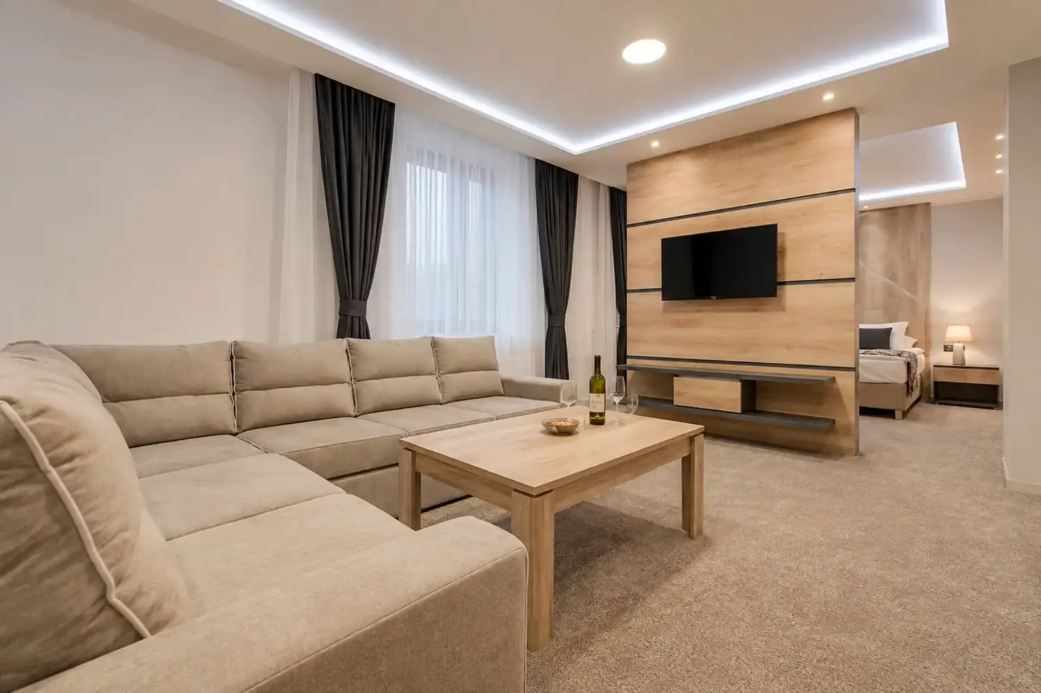 Suite living room with a large corner sofa, timber media wall and bedroom beyond