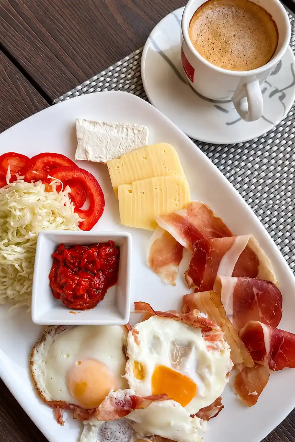 Breakfast plate with fried eggs, cured ham, cheese, cabbage salad, ajvar and coffee